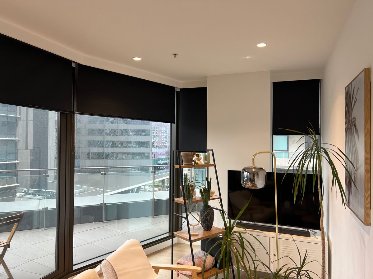 Black roller blinds on the corner windows of an apartment living room overlooking a balcony